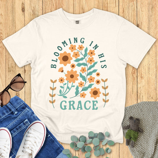 White t-shirt with floral design and text 'Blooming in His Grace' on a wooden surface with accessories.