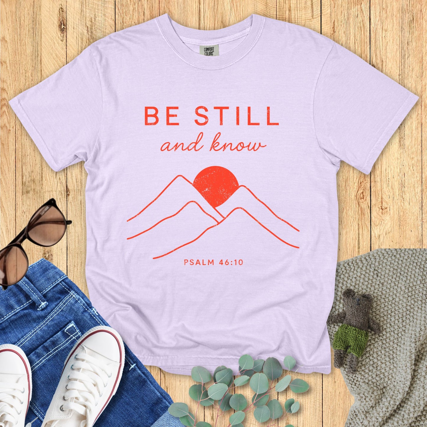Light purple t-shirt with red mountain and sun design, 'Be Still and Know' text on a wooden surface with accessories.