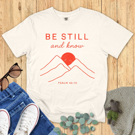 White t-shirt with red mountain design and 'Be Still and Know' text on a wooden surface with jeans and sneakers.