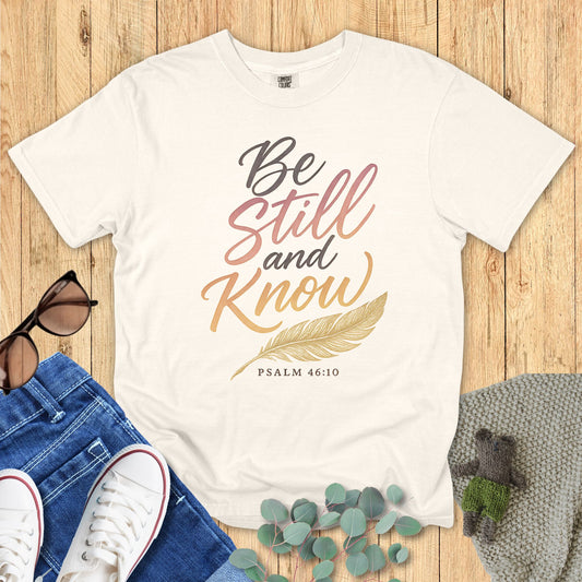 Ivory t-shirt with 'Be Still and Know' text on a wooden surface with accessories.
