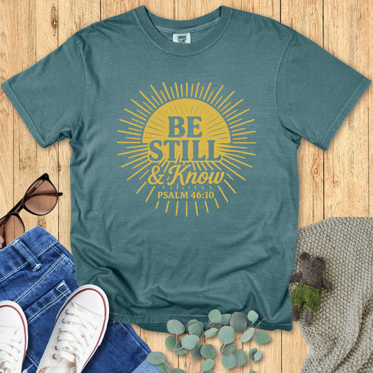 Teal t-shirt with 'Be Still & Know' design on a wooden surface with accessories.