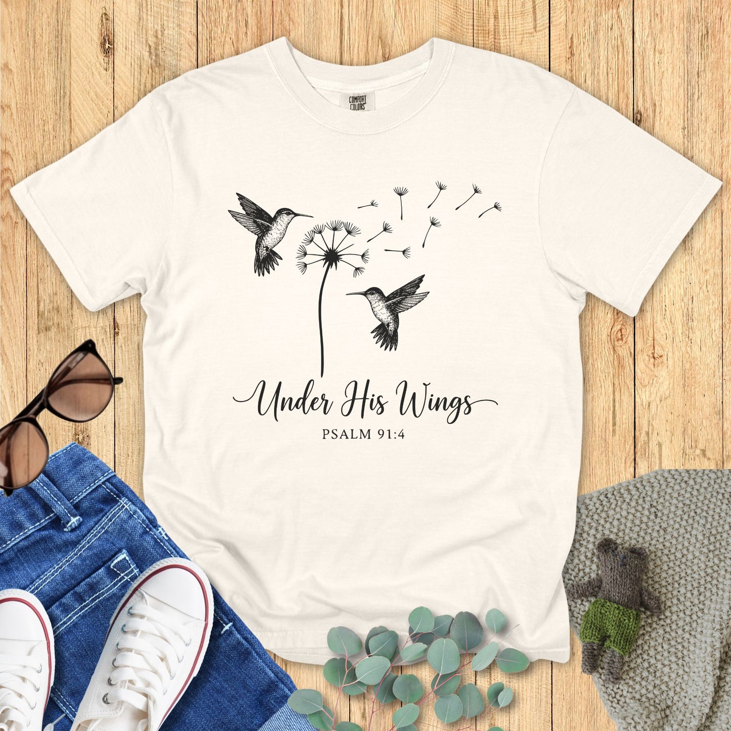 White t-shirt with hummingbird and dandelion design on a wooden surface with accessories.
