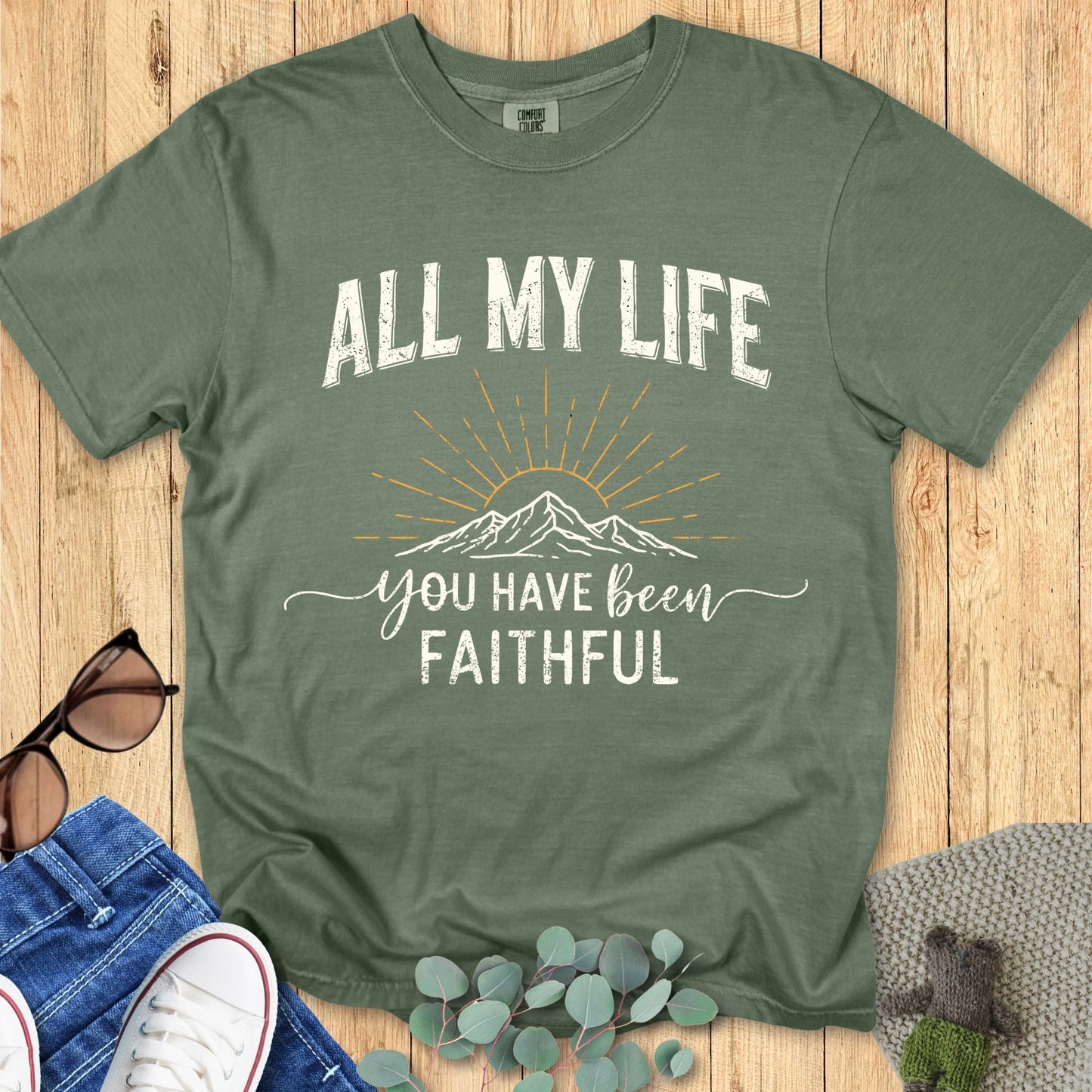 A moss green Comfort Colors T-shirt displayed on a wooden surface. The graphic design features “ALL MY LIFE” in distressed white uppercase letters above a black line art mountain with golden sun rays. Below, the phrase “you have been faithful” is written in script and block font. The flat lay styling includes denim jeans, white sneakers, sunglasses, and eucalyptus leave