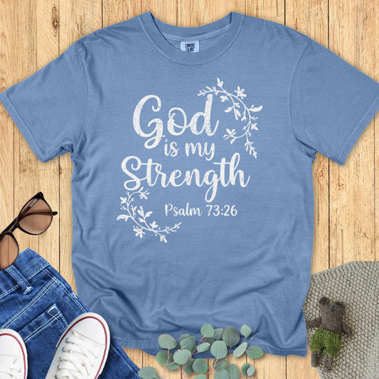 Light blue Comfort Colors T-shirt with distressed white text that reads “God is my Strength – Psalm 73:26,” decorated with floral vine graphics. Flat lay setup with sunglasses, denim jeans, white sneakers, and greenery on a wooden surface.