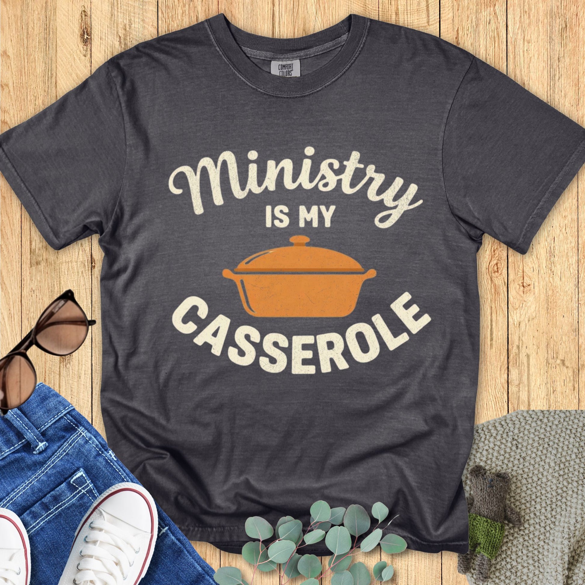 Gray t-shirt with 'Ministry is my Casserole' design on a wooden surface with sunglasses and jeans.