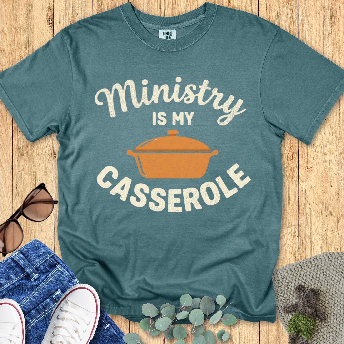 Teal t-shirt with 'Ministry is my Casserole' graphic on a wooden surface with sunglasses and jeans.