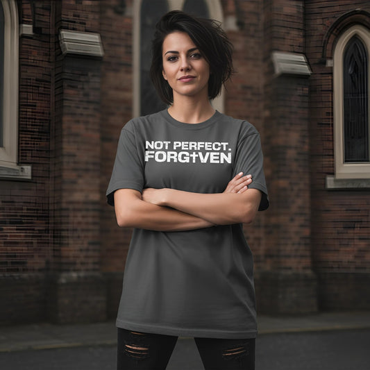 Person wearing a gray t-shirt with 'NOT PERFECT. FORGOTTEN' text in front of a brick building.