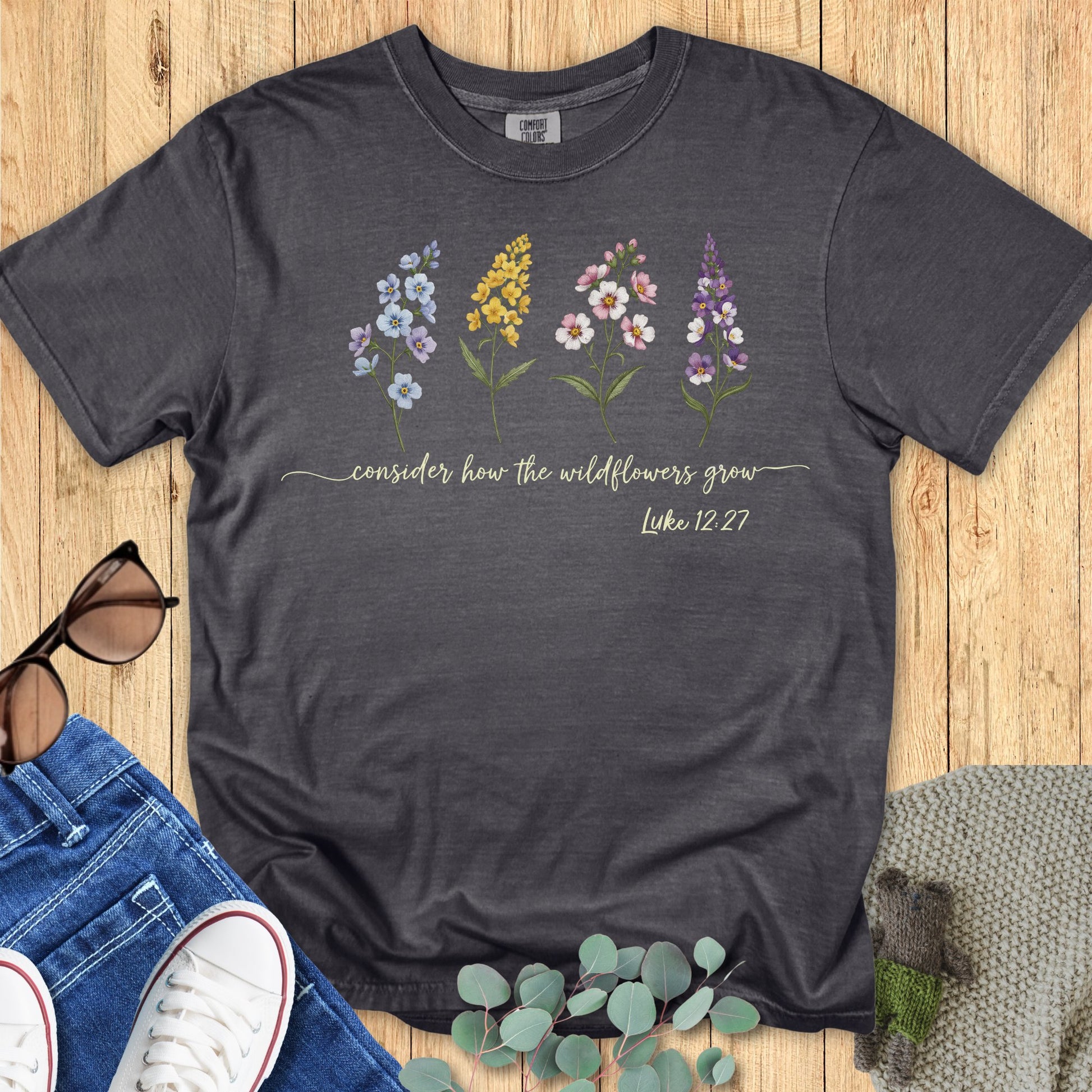 Comfort Colors 1717 Christian women t-shirt in pepper gray flat lay. Design features four colorful wildflowers with the verse “Consider how the wildflowers grow – Luke 12:27” in script fon
