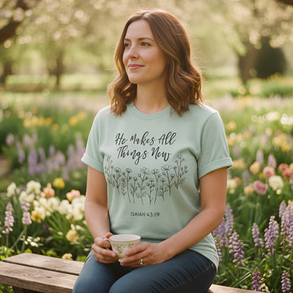 Woman sitting in a flower garden holding a teacup, wearing a bay green Comfort Colors 1717 Christian women t-shirt. The shirt design reads “He Makes All Things New – Isaiah 43:19” with hand-drawn floral illustrations