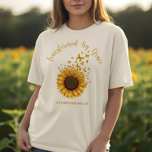 Woman standing in a sunflower field wearing an ivory Comfort Colors 1717 Christian women t-shirt. Shirt design shows a sunflower with butterflies and the words “Transformed by Grace – 2 Corinthians 5:17