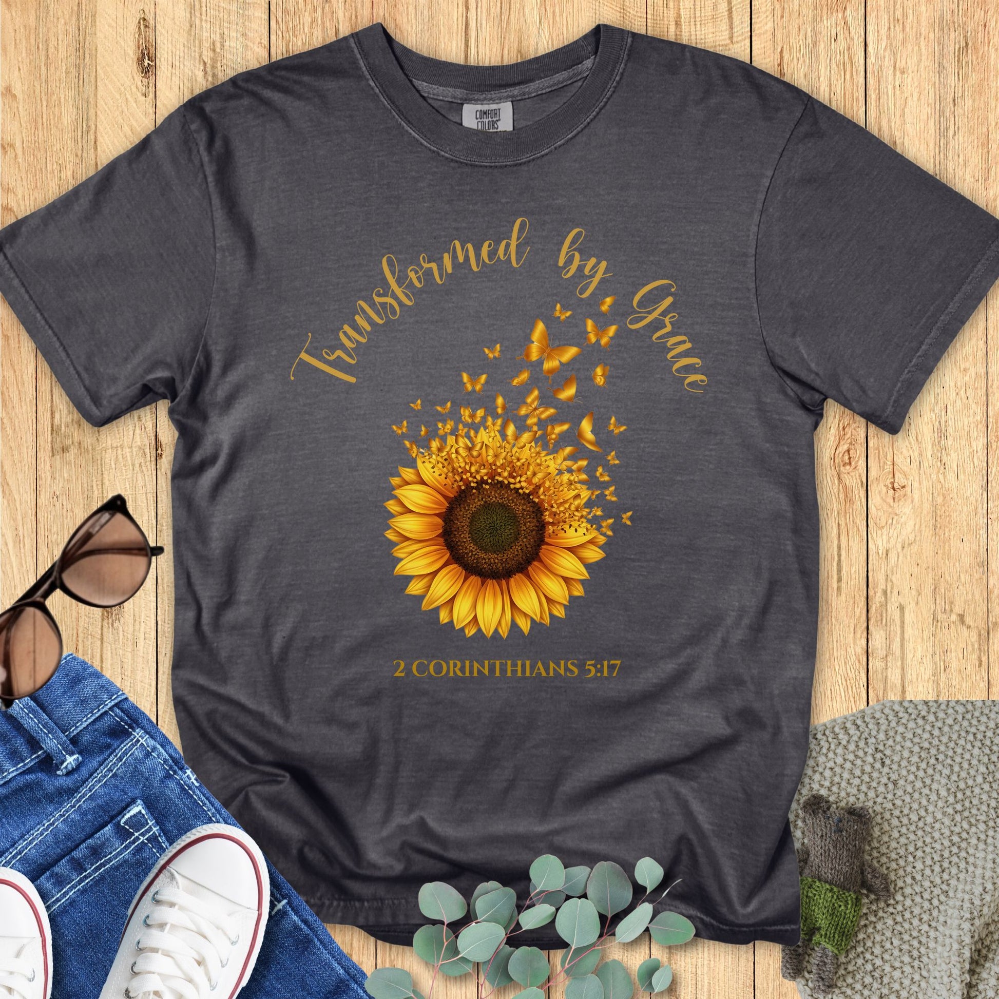 Comfort Colors 1717 Christian women t-shirt in pepper gray flat lay. Design features a sunflower with golden butterflies and the phrase “Transformed by Grace – 2 Corinthians 5:17