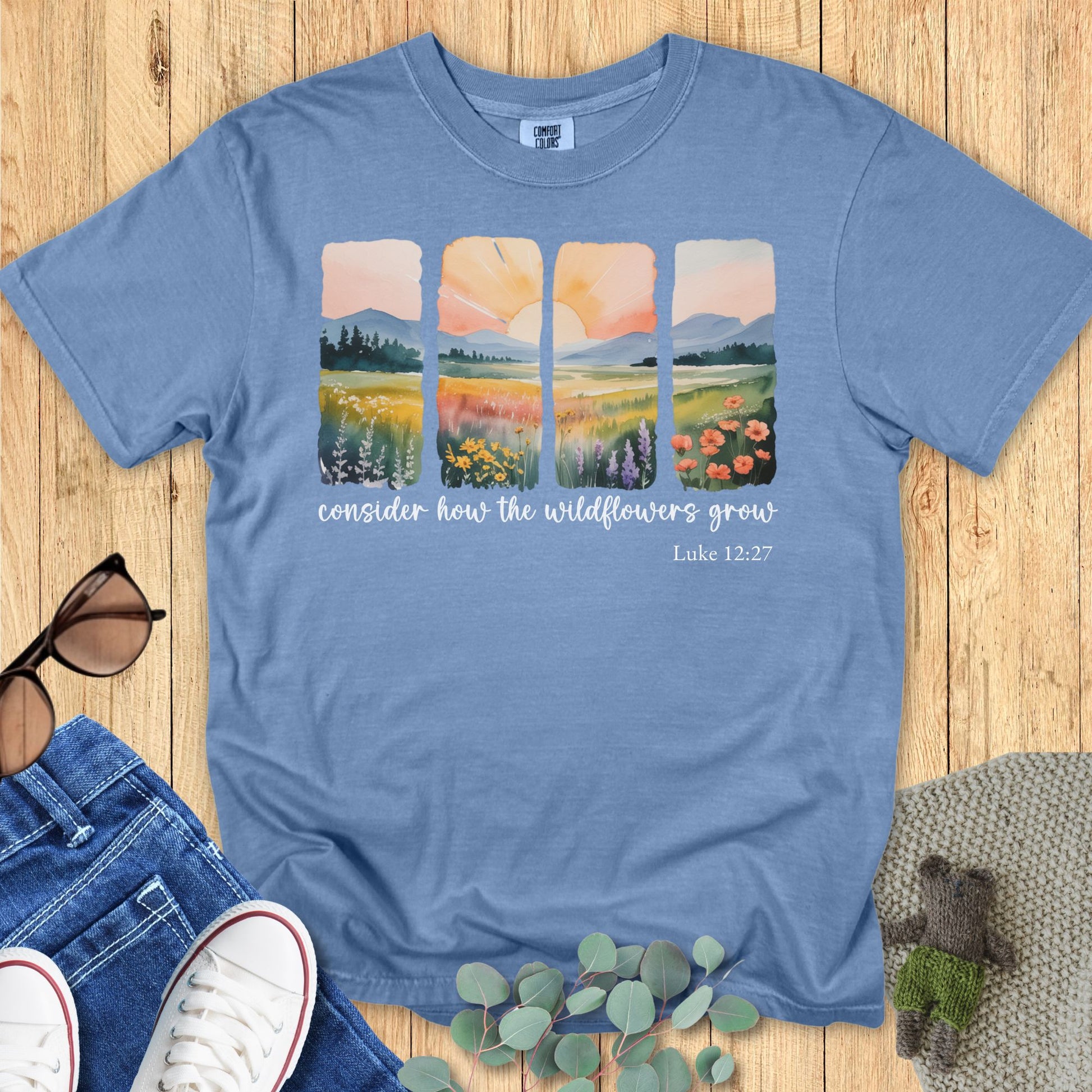 Blue Jean Comfort Colors T-shirt featuring watercolor-style four-panel wildflower landscape and sunrise design, scripture “Consider how the wildflowers grow – Luke 12:27.” Flat lay with jeans, sneakers, sunglasses, and eucalyptus on wooden background