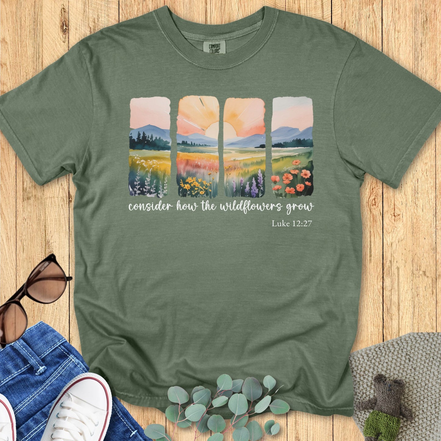 Moss green Comfort Colors T-shirt featuring a watercolor-style four-panel landscape with wildflowers, mountains, and sunrise, with the text “Consider how the wildflowers grow – Luke 12:27.” Flat lay with jeans, sneakers, sunglasses, and eucalyptus on wooden background