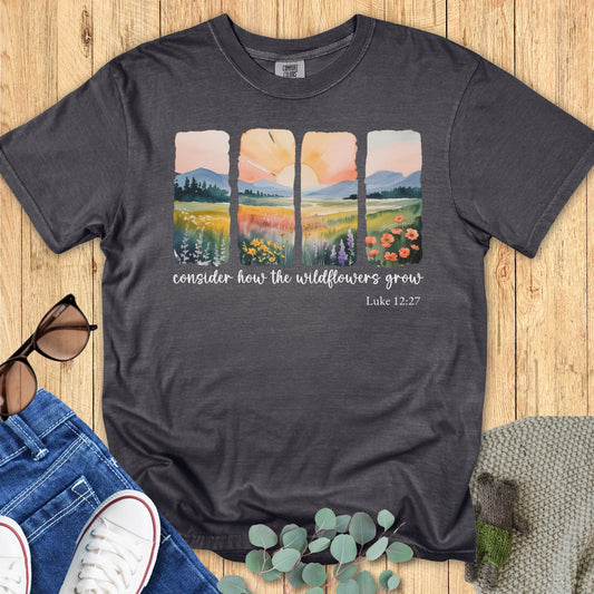 harcoal pepper Comfort Colors T-shirt with a watercolor-style four-panel wildflower landscape and sunrise design, scripture text “Consider how the wildflowers grow – Luke 12:27.” Flat lay with jeans, sneakers, sunglasses, and eucalyptus on wooden background