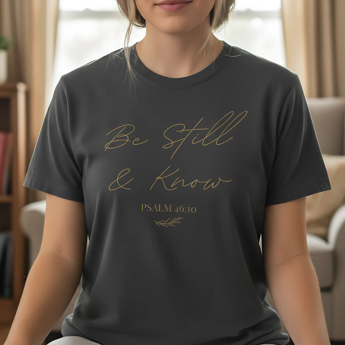 Woman wearing a dark gray T-shirt with the phrase "Be Still & Know" and Psalm 46:10 in script, sitting peacefully indoors, suggesting calm and meditation