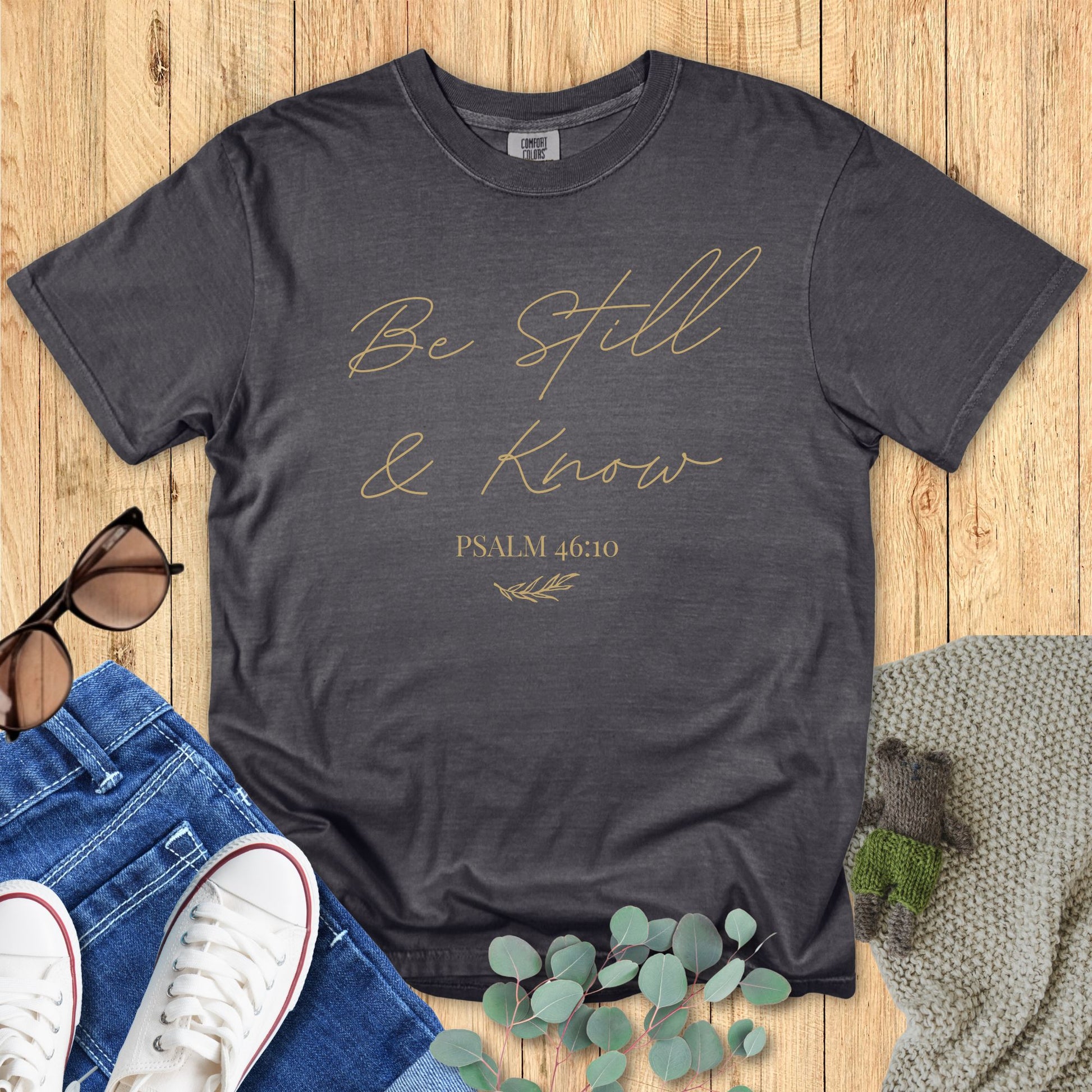 Dark pepper gray Comfort Colors T-shirt with minimalist script text "Be Still & Know" and Psalm 46:10, styled flat with denim, sunglasses, and a knitted accessory.