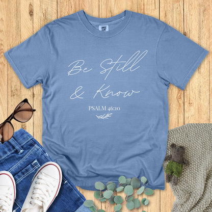 Blue jean Comfort Colors T-shirt with the phrase "Be Still & Know" and Psalm 46:10 in delicate script font, styled with jeans, sunglasses, and greenery on a wooden background