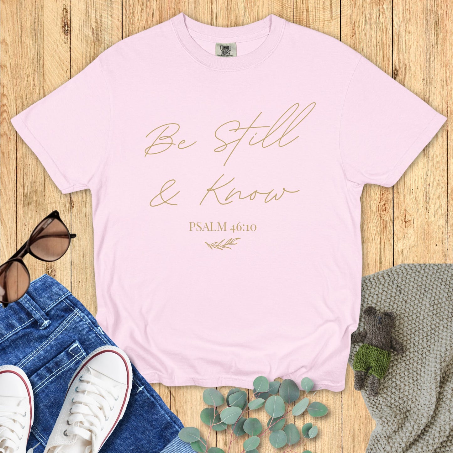 Blossom pink Comfort Colors T-shirt with elegant script text "Be Still & Know" and Psalm 46:10, arranged with jeans, sunglasses, and greenery on a rustic wood backdrop.