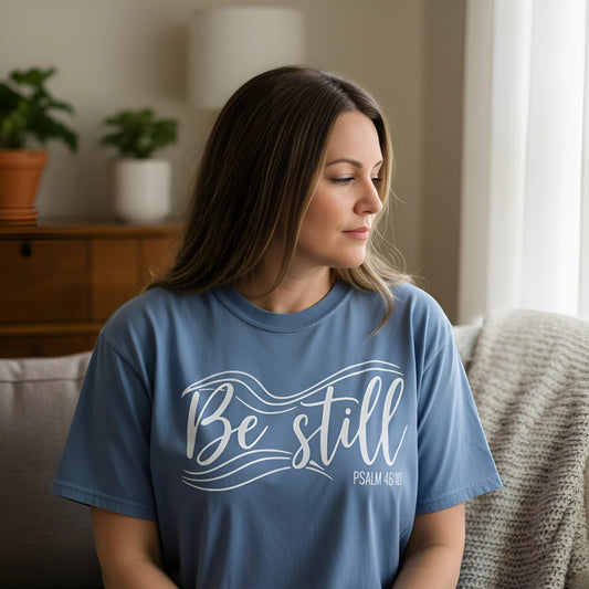 Woman sitting indoors wearing a blue jean Christian T-shirt with the words “Be Still” in bold white script, accented with flowing wave lines and “Psalm 46:10.” She is seated near a window, looking out peacefully with plants in the background.