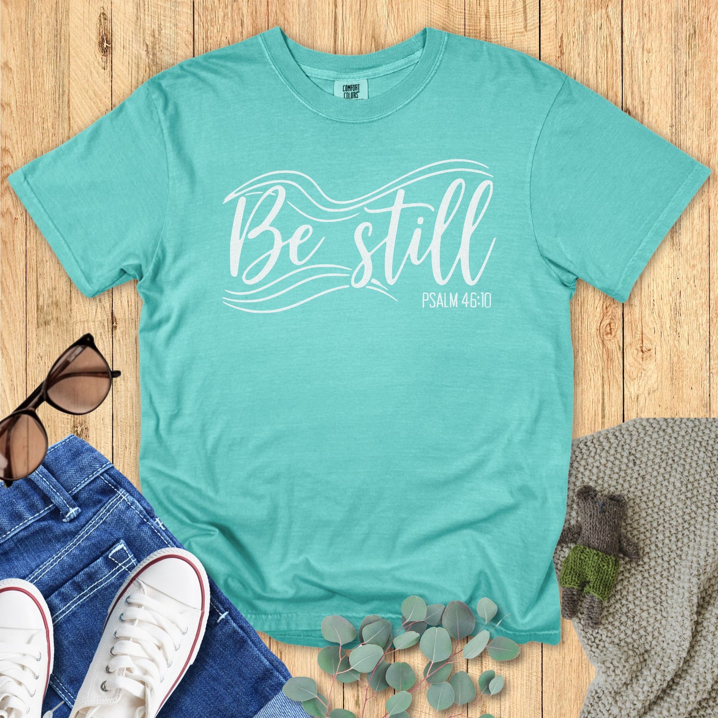 Seafoam green Christian T-shirt flat lay with the phrase “Be Still” in elegant white lettering and flowing wave graphics, alongside “Psalm 46:10.” Styled with sunglasses, jeans, sneakers, eucalyptus leaves, and a knitted blanket on a wooden background