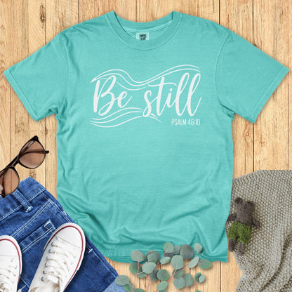 Seafoam green Christian T-shirt flat lay with the phrase “Be Still” in elegant white lettering and flowing wave graphics, alongside “Psalm 46:10.” Styled with sunglasses, jeans, sneakers, eucalyptus leaves, and a knitted blanket on a wooden background