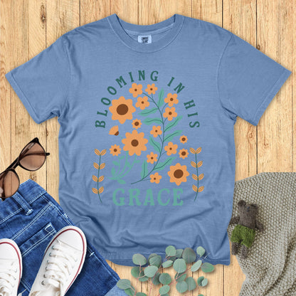 Comfort Colors 1717 Christian women t-shirt in blue, printed with “Blooming in His Grace” in teal text surrounded by orange floral artwork. Styled flat with jeans, sneakers, sunglasses, eucalyptus sprigs, and a knitted bear on a wooden background.