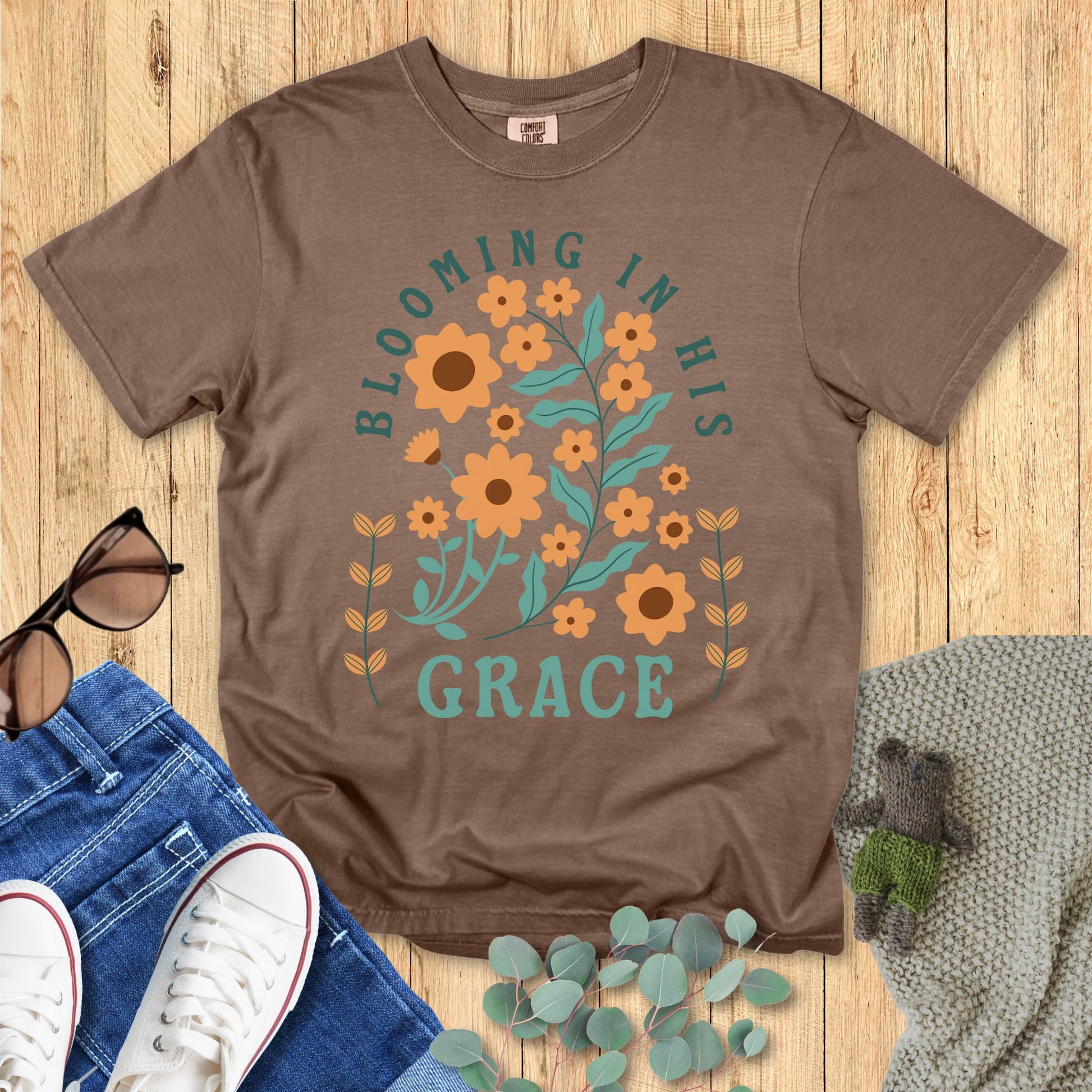 Comfort Colors 1717 Christian women t-shirt in brown, featuring the phrase “Blooming in His Grace” with orange flowers and green leaves in a faith-inspired floral design. Styled with jeans, sneakers, sunglasses, and eucalyptus on a wooden background