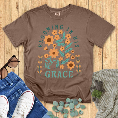 Comfort Colors 1717 Christian women t-shirt in brown, featuring the phrase “Blooming in His Grace” with orange flowers and green leaves in a faith-inspired floral design. Styled with jeans, sneakers, sunglasses, and eucalyptus on a wooden background