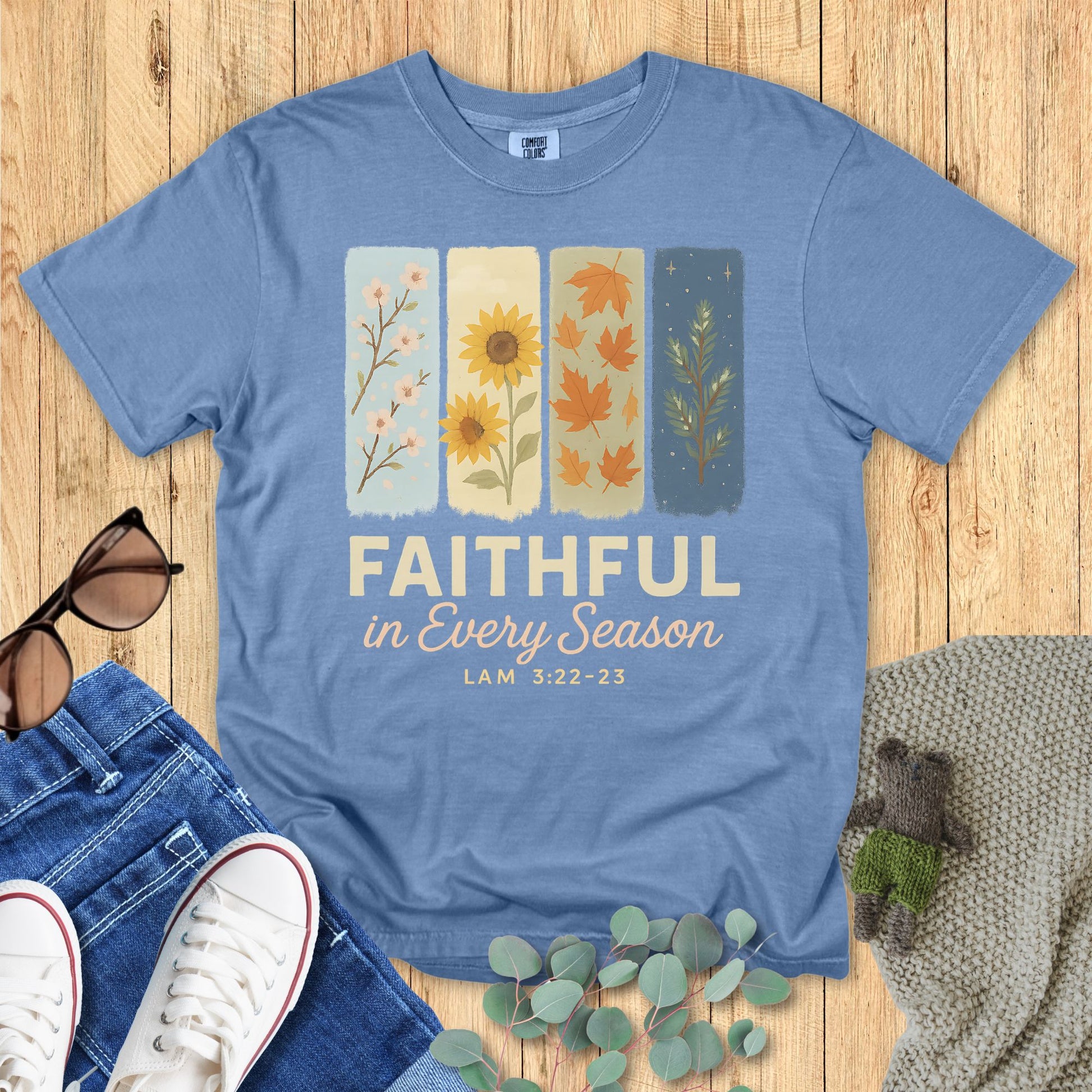 Flat lay of Comfort Colors 1717 Blue Jean Christian women’s t-shirt. Shirt design includes four watercolor-style panels showing flowers and leaves across the seasons with scripture text “Faithful in Every Season – Lam 3:22-23.” Surrounded by casual lifestyle props like sunglasses, jeans, and greenery