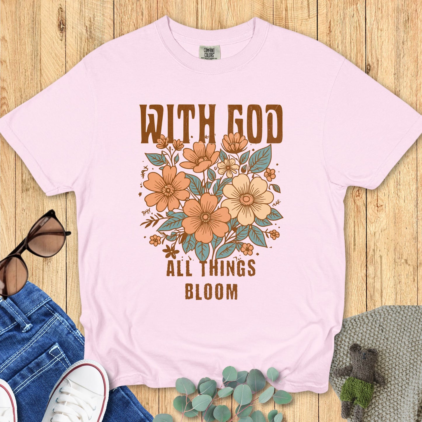 Christian women’s Comfort Colors 1717 t-shirt in Blossom pink with vintage-style florals and text ‘With God All Things Bloom.’ Flat lay styled with jeans, sunglasses, and cozy accents