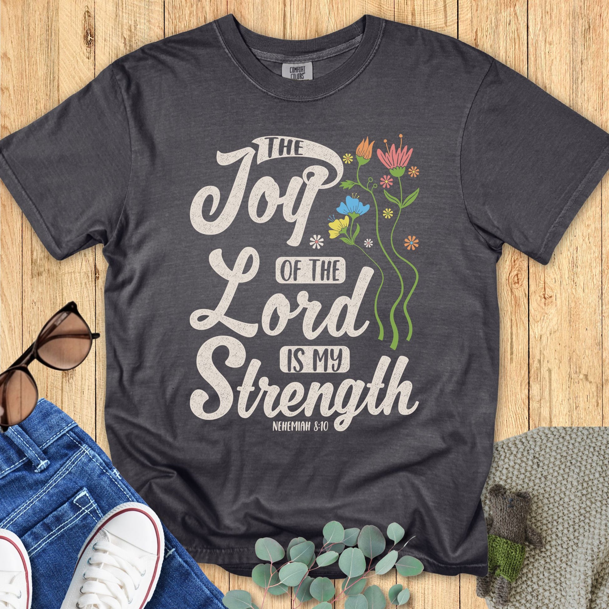 Comfort Colors 1717 Christian women’s t-shirt in pepper gray, styled flat with jeans, sunglasses, and greenery. Design features colorful flowers beside the Bible verse “The Joy of the Lord is My Strength – Nehemiah 8:10