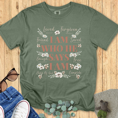 Sage green Christian women’s t-shirt featuring the affirmation “I Am Who He Says I Am” with scripture references and soft floral artwork, flat lay styled with jeans, sneakers, sunglasses, and eucalyptus for a boutique aesthetic