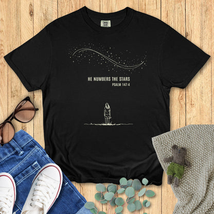 Black Comfort Colors 1717 women’s Christian T-shirt laid flat on wood background, decorated with white sneakers, denim jeans, and sunglasses. Design reads “He Numbers the Stars – Psalm 147:4” with a starry night illustration
