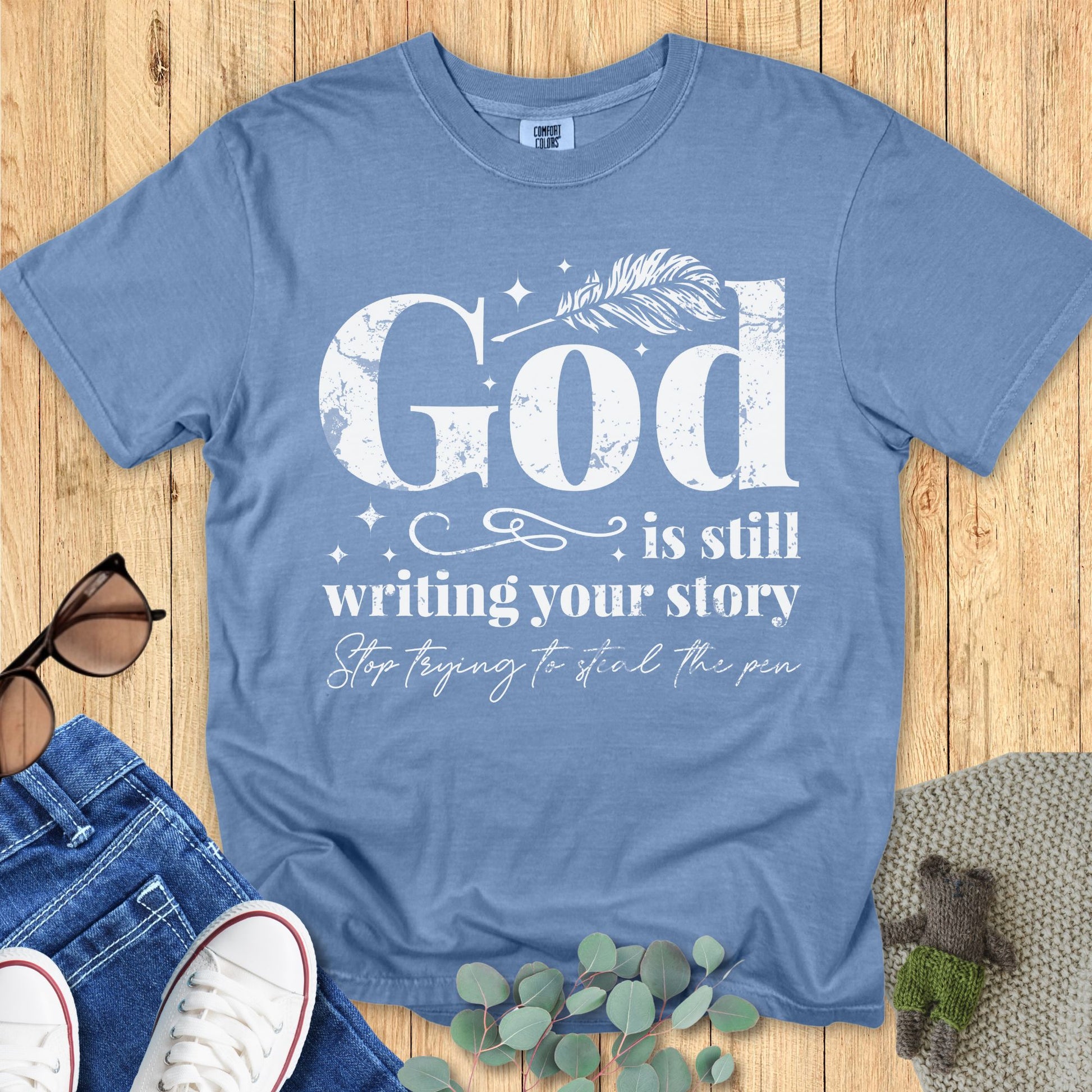 Christian Comfort Colors 1717 T-shirt in blue jean color featuring the phrase “God is still writing your story, stop trying to steal the pen” in distressed white lettering, flat lay with casual accessories