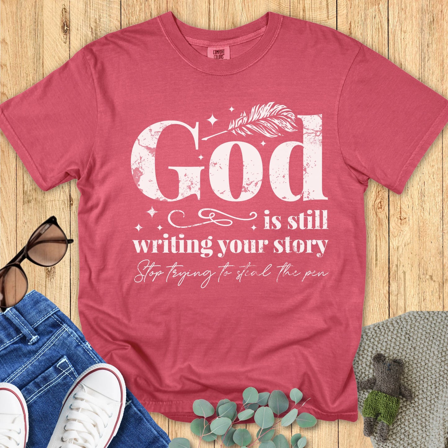 Faith-based Comfort Colors 1717 T-shirt in crimson with distressed white typography that says “God is still writing your story, stop trying to steal the pen,” displayed flat on wood background with jeans and sunglasse