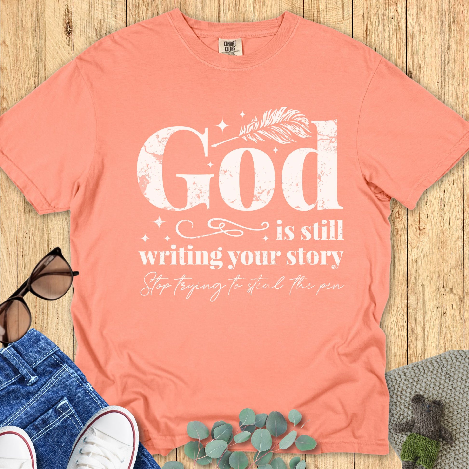 Christian women’s Comfort Colors 1717 T-shirt in terracotta with white text design that reads “God is still writing your story, stop trying to steal the pen,” styled with sunglasses, jeans, and sneakers on a wooden background