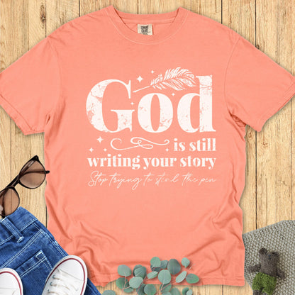 Christian women’s Comfort Colors 1717 T-shirt in terracotta with white text design that reads “God is still writing your story, stop trying to steal the pen,” styled with sunglasses, jeans, and sneakers on a wooden background