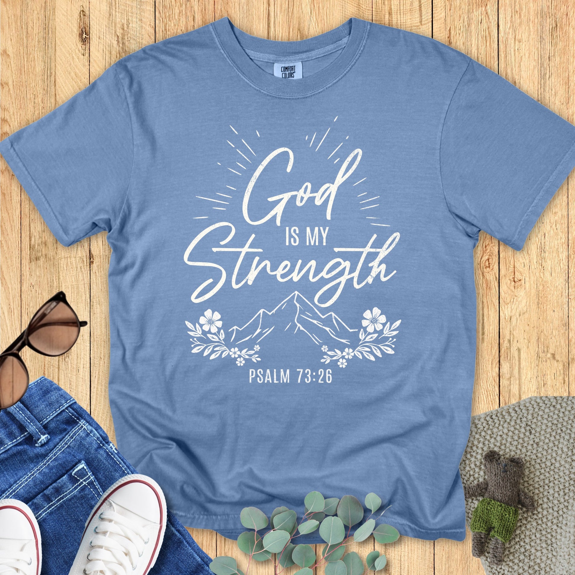 Comfort Colors 1717 blue jean T-shirt with white lettering “God is my Strength – Psalm 73:26” and decorative mountain and floral artwork. Displayed flat on wooden surface with lifestyle styling.