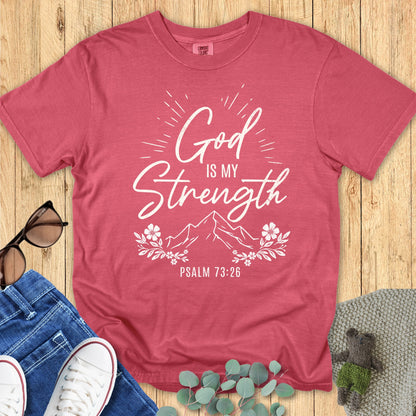 Comfort Colors 1717 crimson unisex T-shirt with white Christian design reading “God is my Strength – Psalm 73:26” in script and bold fonts, with mountain illustration, sunburst rays, and floral accents. Flat lay styled with sunglasses, denim jeans, sneakers, and greenery on a wooden background
