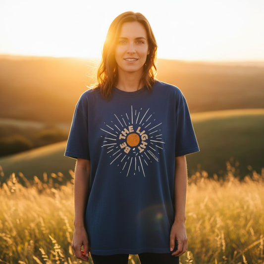 Woman standing in a golden field at sunset wearing a midnight blue Comfort Colors 1717 Christian t-shirt with a radiant sunburst graphic. The shirt reads “Be The Light” with Matthew 5:14, symbolizing faith and encouragement