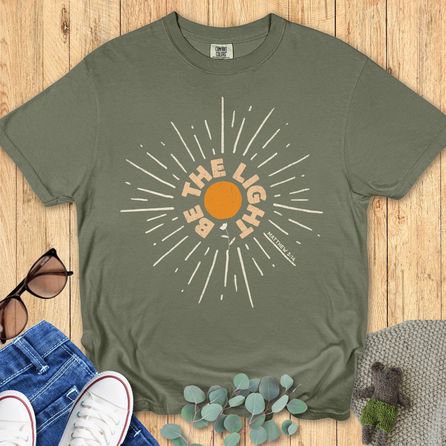 Sage green Comfort Colors 1717 Christian t-shirt laid flat on a wooden background. The print displays an orange sunburst with bold lettering: “Be The Light – Matthew 5:14,” emphasizing a message of hope and faith.