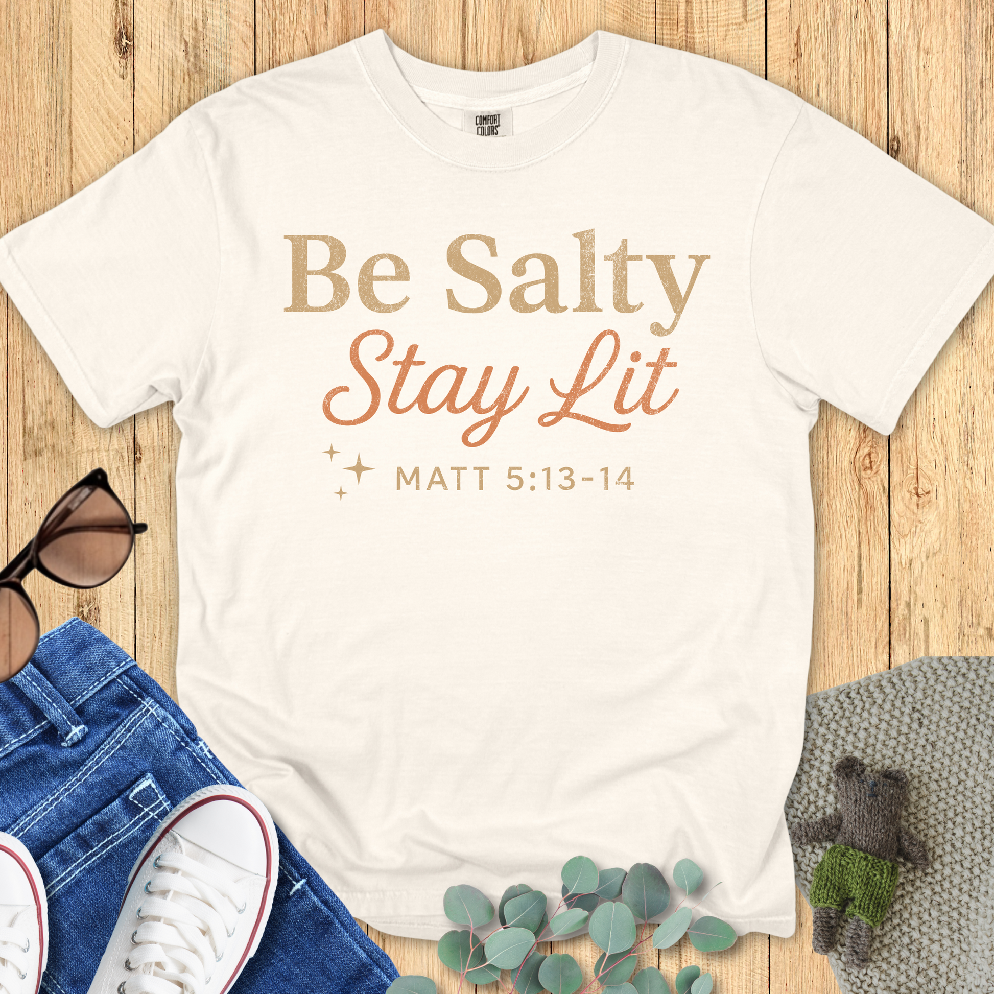 Comfort Colors 1717 Christian women’s T-shirt in ivory with the phrase “Be Salty Stay Lit – Matt 5:13-14” in bold and script fonts, styled with casual flat lay including sunglasses, jeans, and sneakers