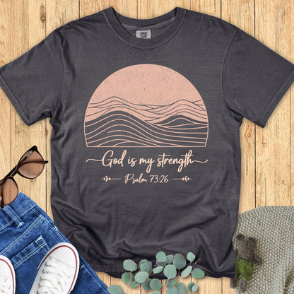 Pepper gray Comfort Colors 1717 Christian t-shirt featuring pink sun and wave artwork with the verse 'God is my strength – Psalm 73:26,' displayed with jeans, white sneakers, and sunglasses on wooden surface