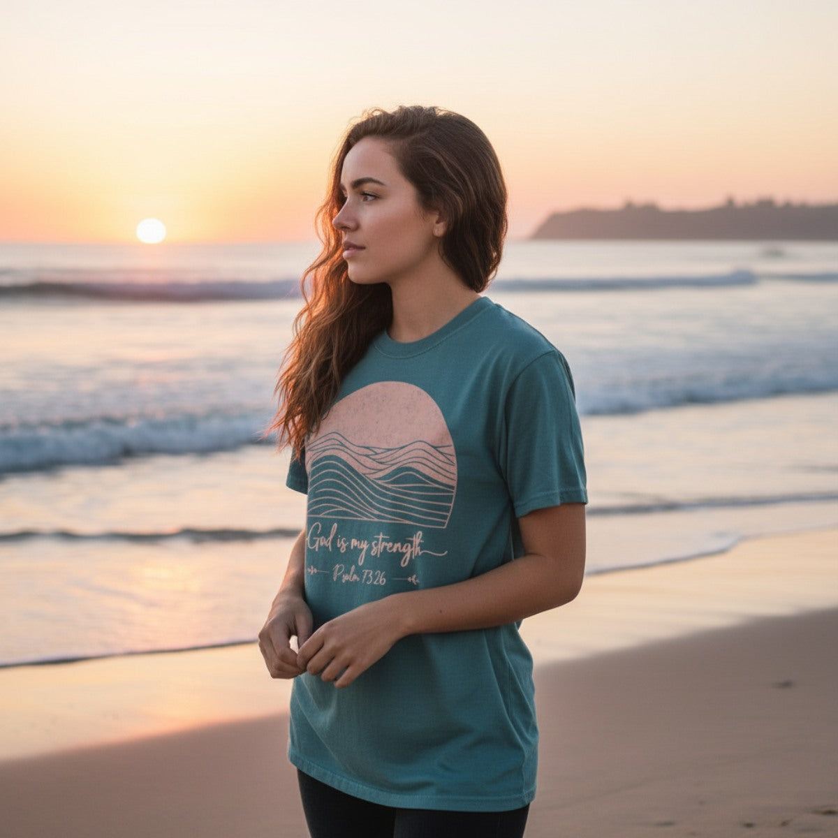 Woman standing on the beach at sunset wearing a teal Comfort Colors 1717 Christian t-shirt with the words 'God is my strength – Psalm 73:26' and a minimalist pink sun and wave design