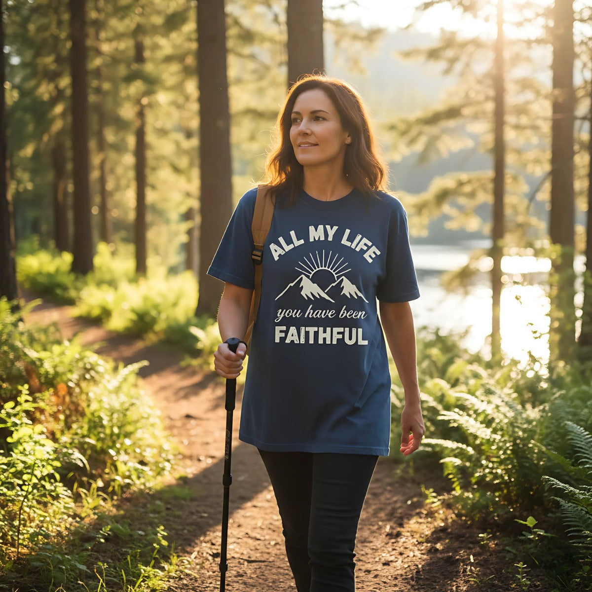 Christian woman hiking in the forest near a lake at sunset, wearing a midnight blue Comfort Colors 1717 T-shirt with mountain and sunrise graphic. Shirt design says 'All My Life You Have Been Faithful.