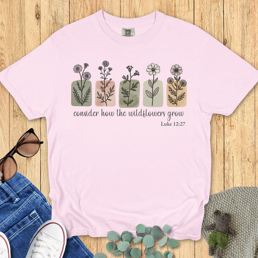 Blossom Comfort Colors 1717 T-shirt mockup – Christian floral graphic tee in soft pink, flat lay with Luke 12:27 design, displayed with jeans and greenery.