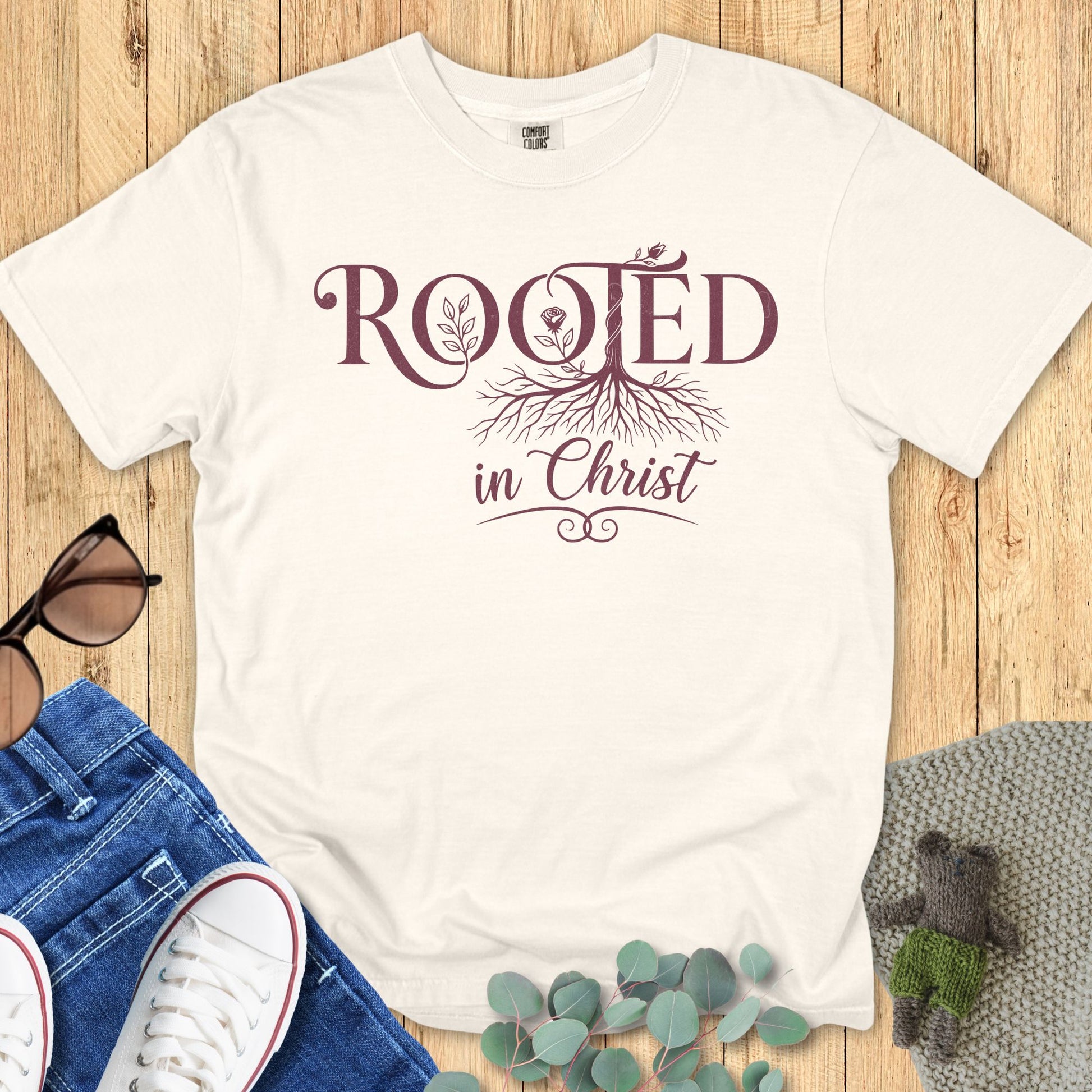 “Ivory Comfort Colors 1717 Christian T-shirt with ‘Rooted in Christ’ design in maroon lettering and tree roots graphic, styled with denim jeans and sneakers on a wooden background.”
