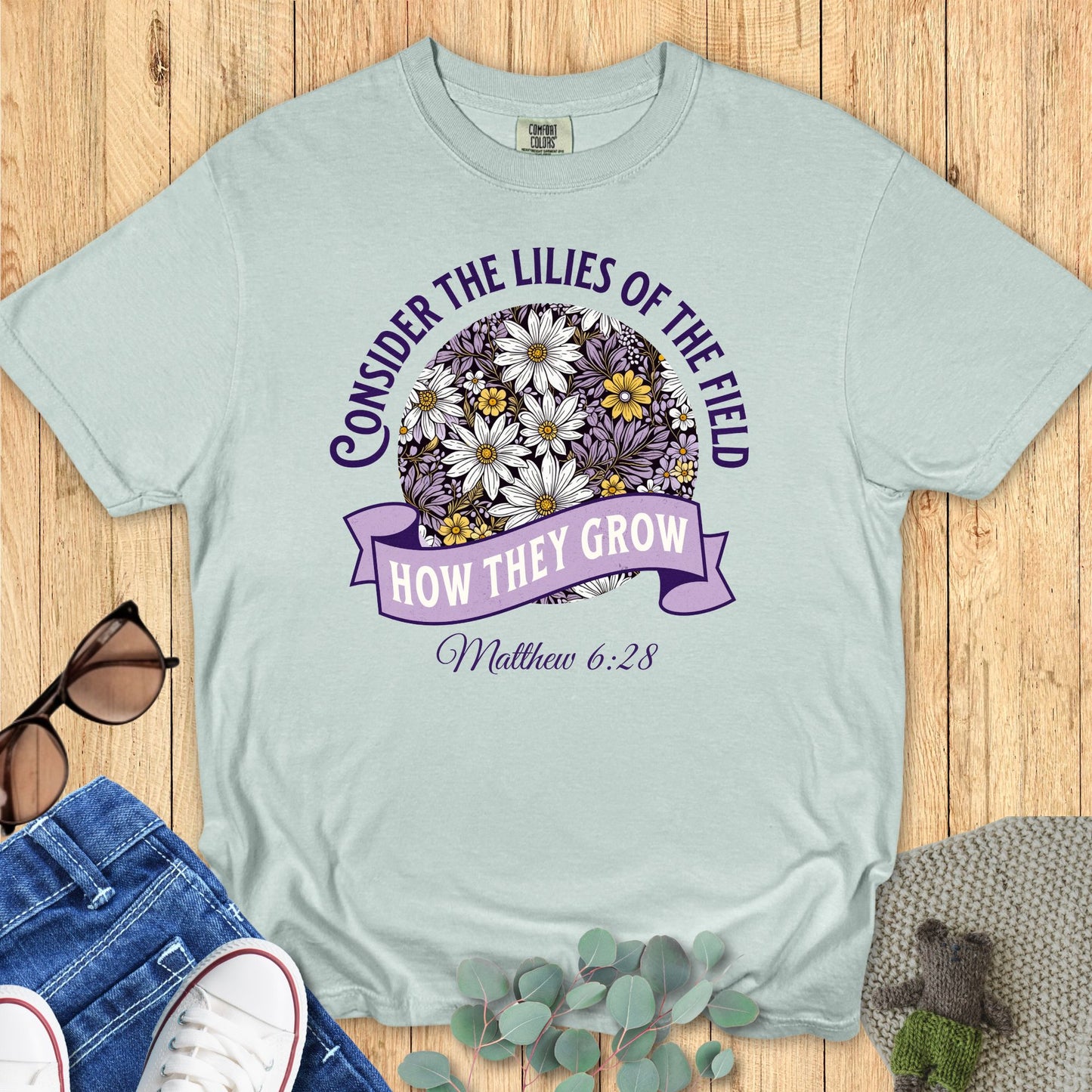 Comfort Colors 1717 Bay blue Christian T-shirt with scripture Matthew 6:28 and daisy illustration in purple, white, and yellow. Styled flat lay on wooden background with jeans and sneakers