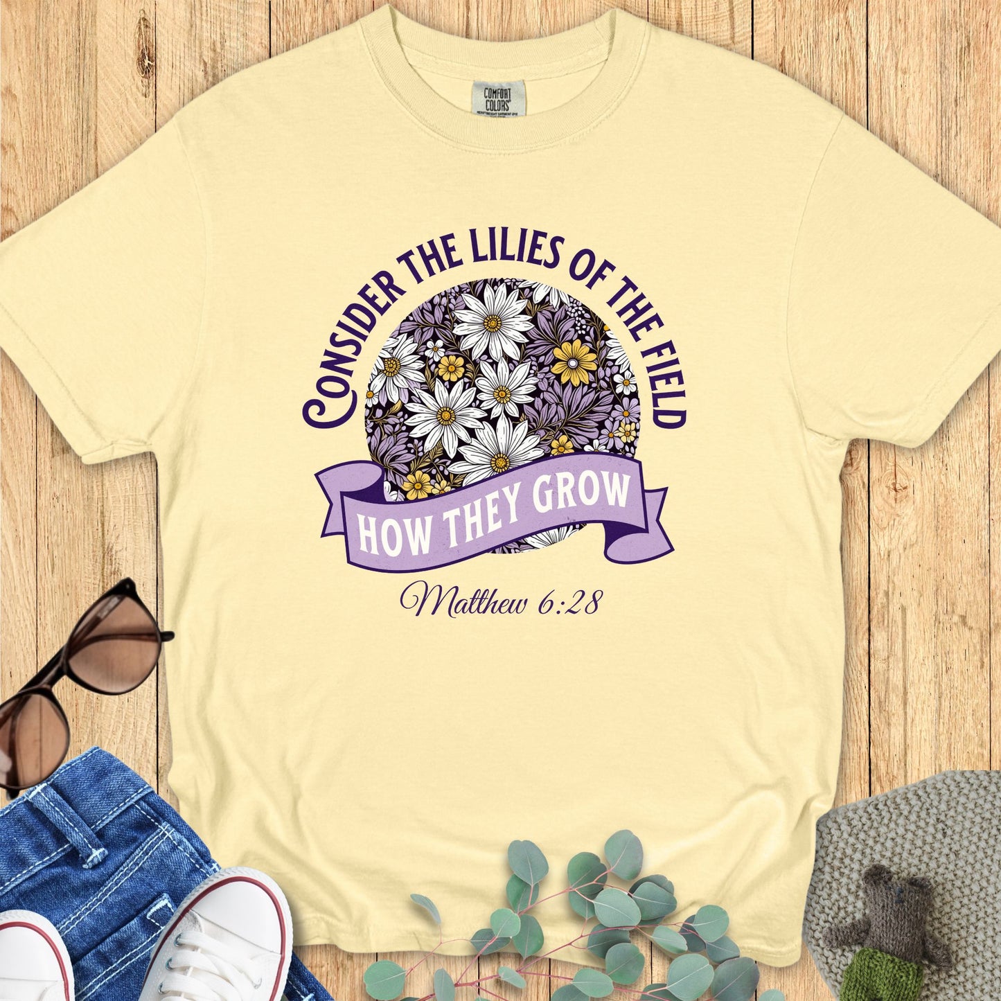 Comfort Colors 1717 Butter yellow Christian T-shirt with purple and white floral graphic. Text reads “Consider the lilies of the field, how they grow – Matthew 6:28.” Styled flat lay with denim, sneakers, and sunglasses.