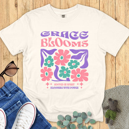 Ivory Comfort Colors 1717 Christian T-shirt with retro floral graphic in pink, green, and purple. Bold text reads ‘Grace Blooms’ with tagline ‘Rooted in Spirit, Blooming with Power.’ Christian apparel flat lay with jeans, sunglasses, and sneakers."